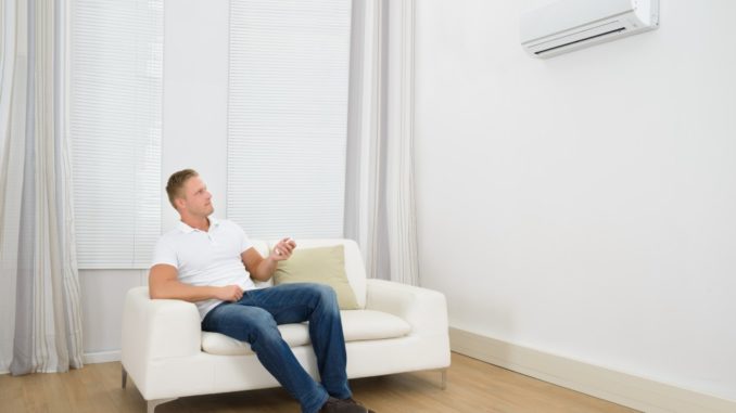 Man Sitting On Sofa Operating Air Conditioner With Remote Controller