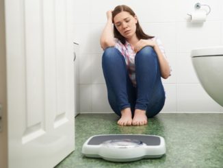 Unhappy Teenage Girl Sitting On Floor Looking At Bathroom Scales