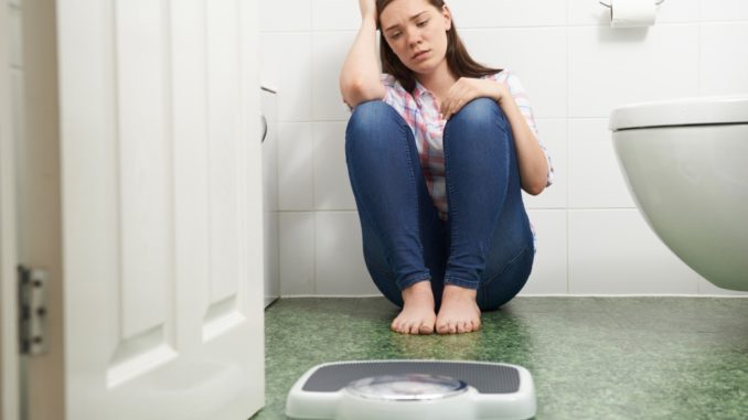 Unhappy Teenage Girl Sitting On Floor Looking At Bathroom Scales