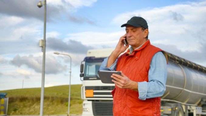 Truck driver talking on the phone while holding a tablet with his truck behind him